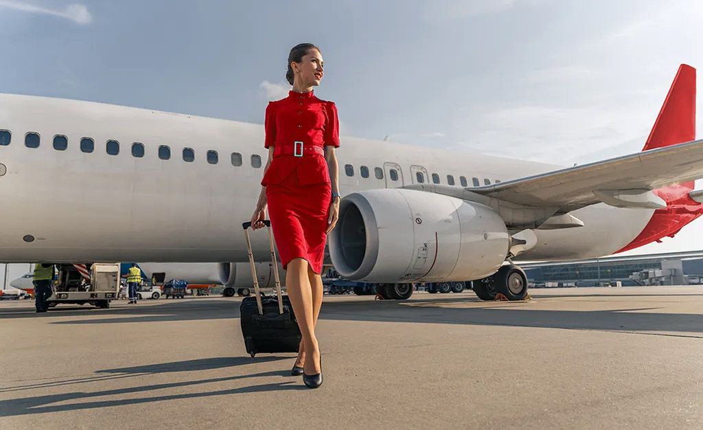 Frankfinn aviation training student in red cabin crew uniform walking with luggage near airplane, showing aviation career lifestyle.